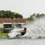 A waterskier in a black vest carving through water, creating a spray, with houses in the background.
