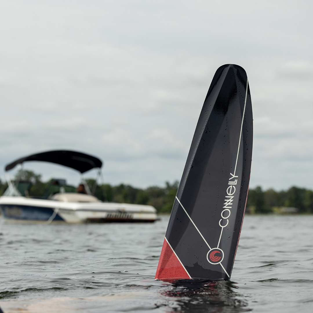 Black Connelly Aspect water ski tip emerging from water, with a boat and trees in the background.