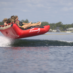 Four riders on Big Easy 4 towable tube, gripping the cushioned backrest as they glide across the water.