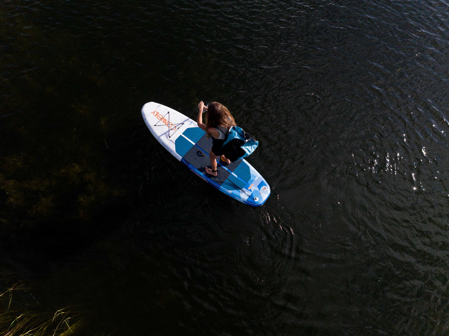 Overhead view of a woman paddling on the Stash iSUP in dark water, carrying the backpack.