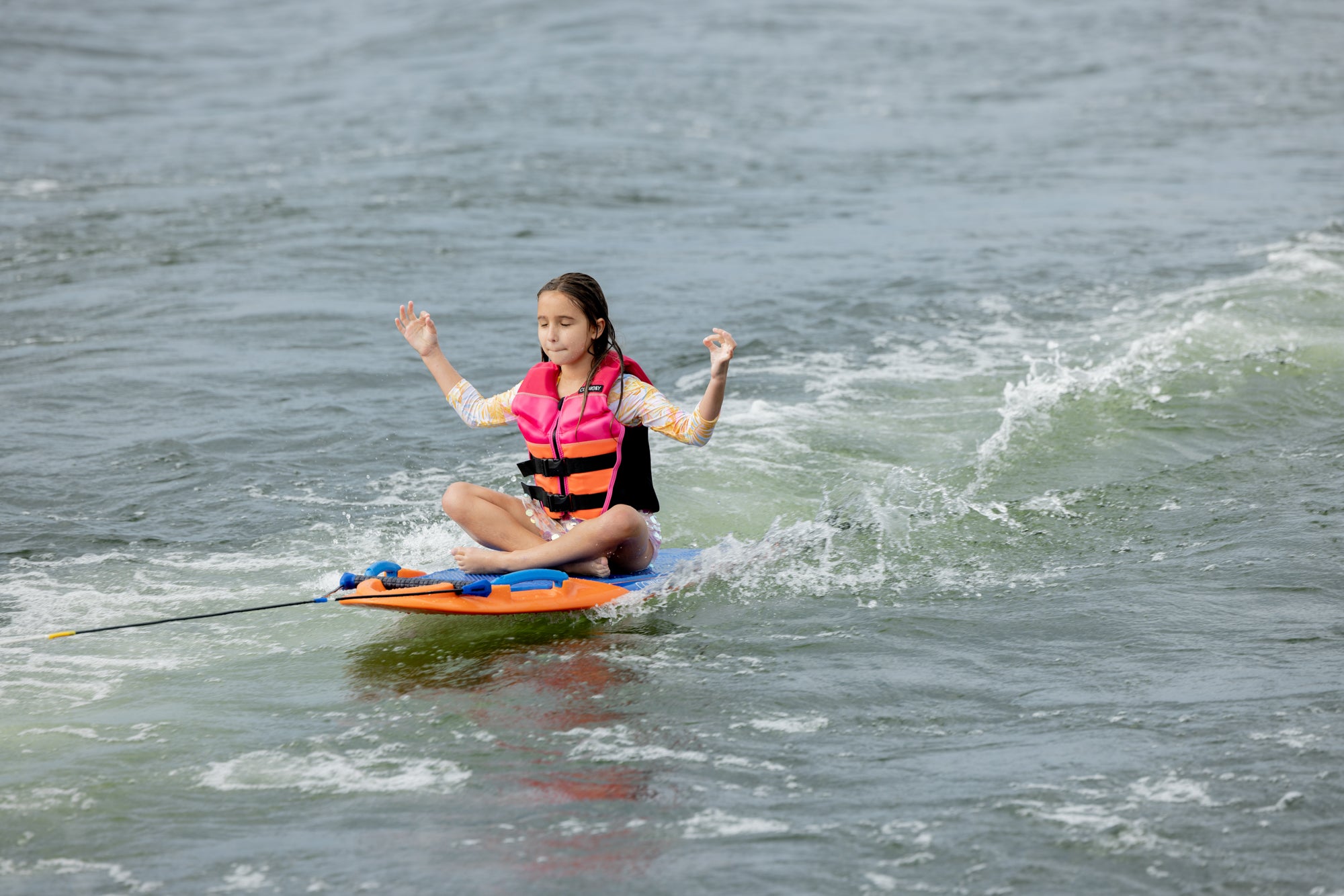 Girl meditating on a kneeboard, being towed on a lake while wearing a pink life jacket.