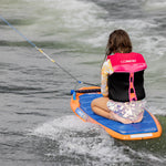Child in pink life jacket kneeling on Wild Thing board being pulled on water.
