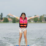 Girl on an orange wakeboard, arms out, wearing a pink Wild Thing life jacket on a lake.