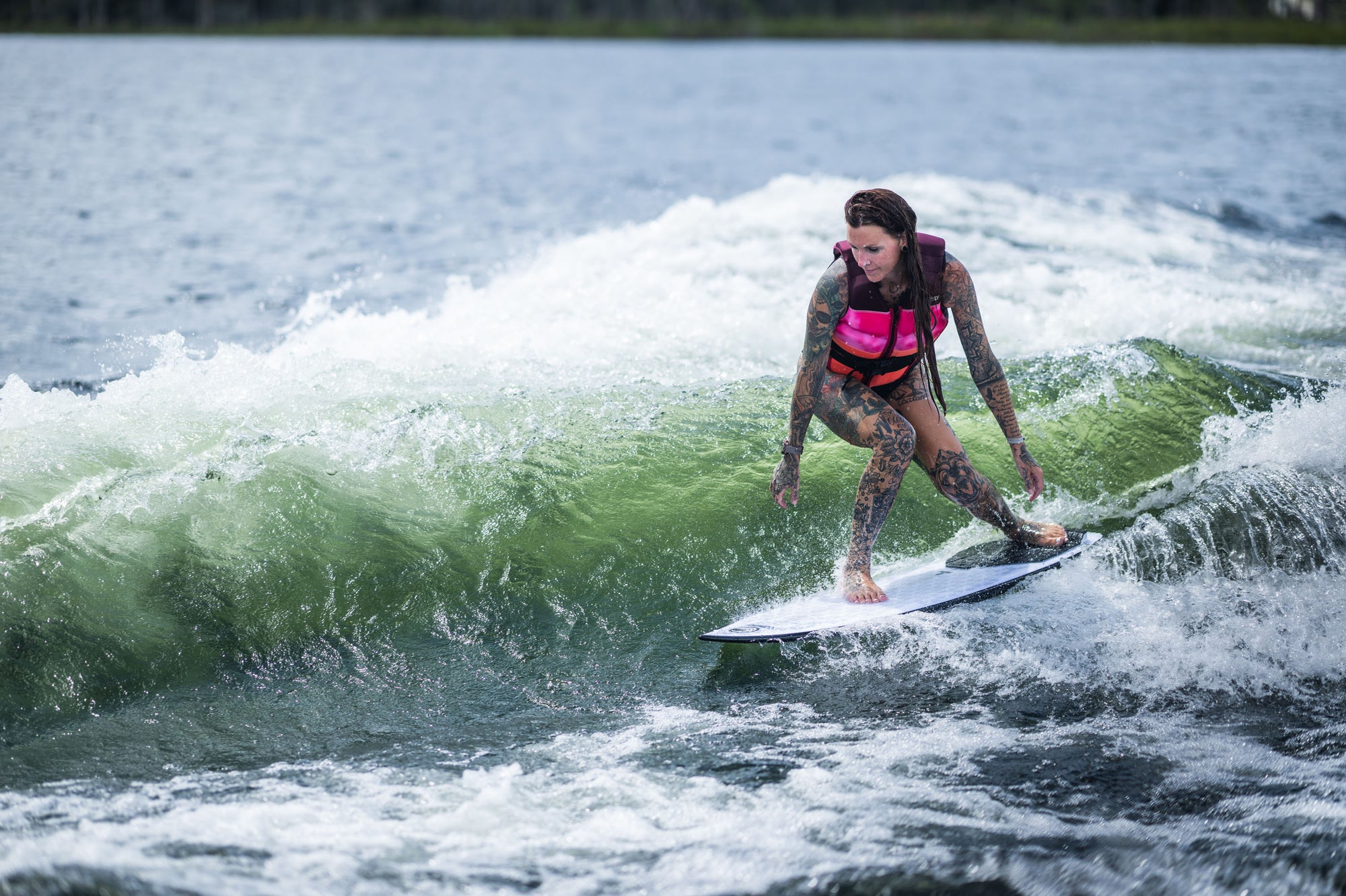Tattooed person in a life vest surfing a wave on a lake.