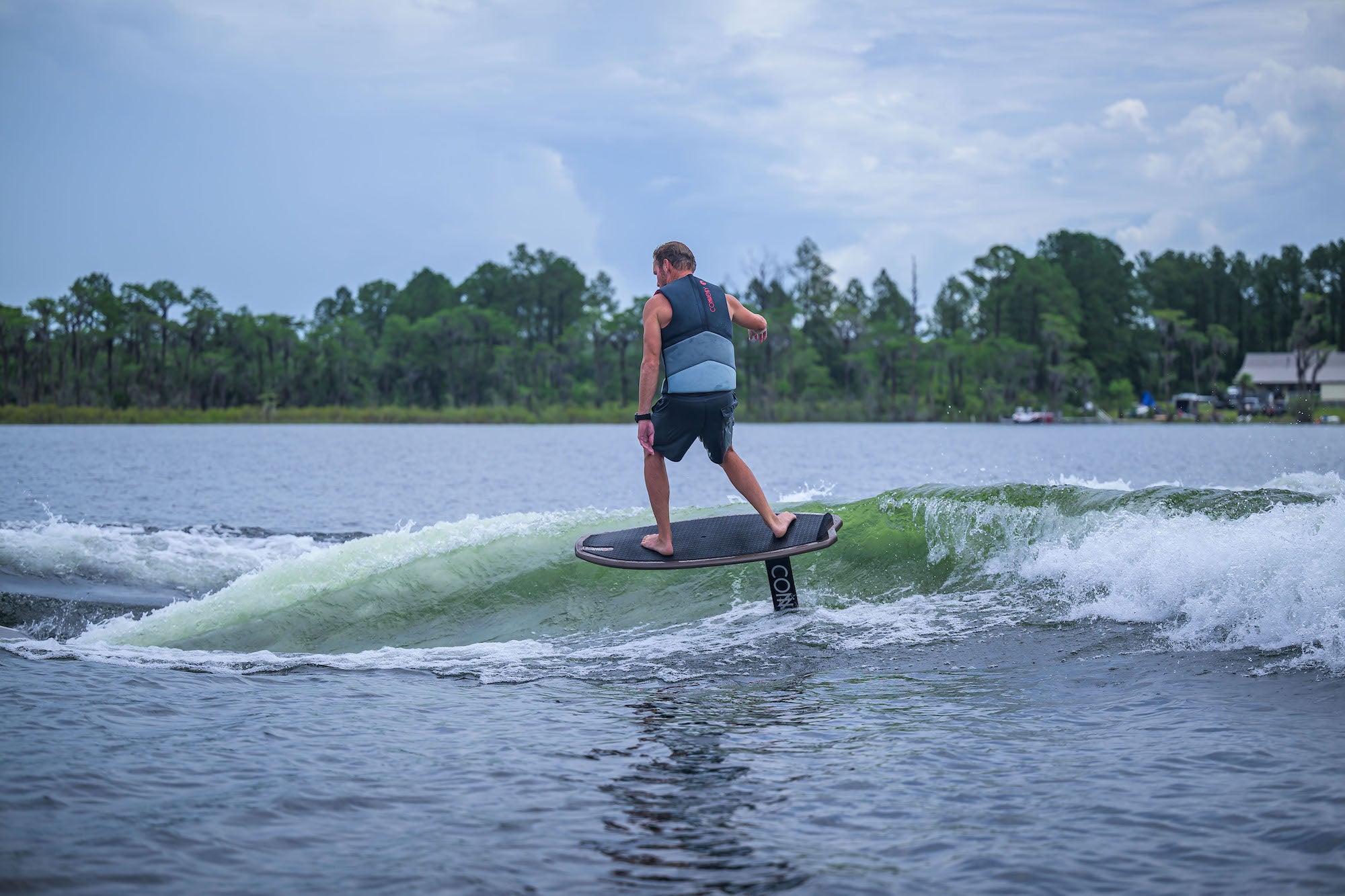 Person riding a wakefoil on a lake with trees and a house in the background.