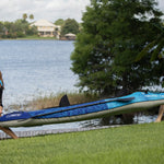 A woman and a man carry the Nautic 11.5 inflatable kayak toward a lake, both wearing life jackets.