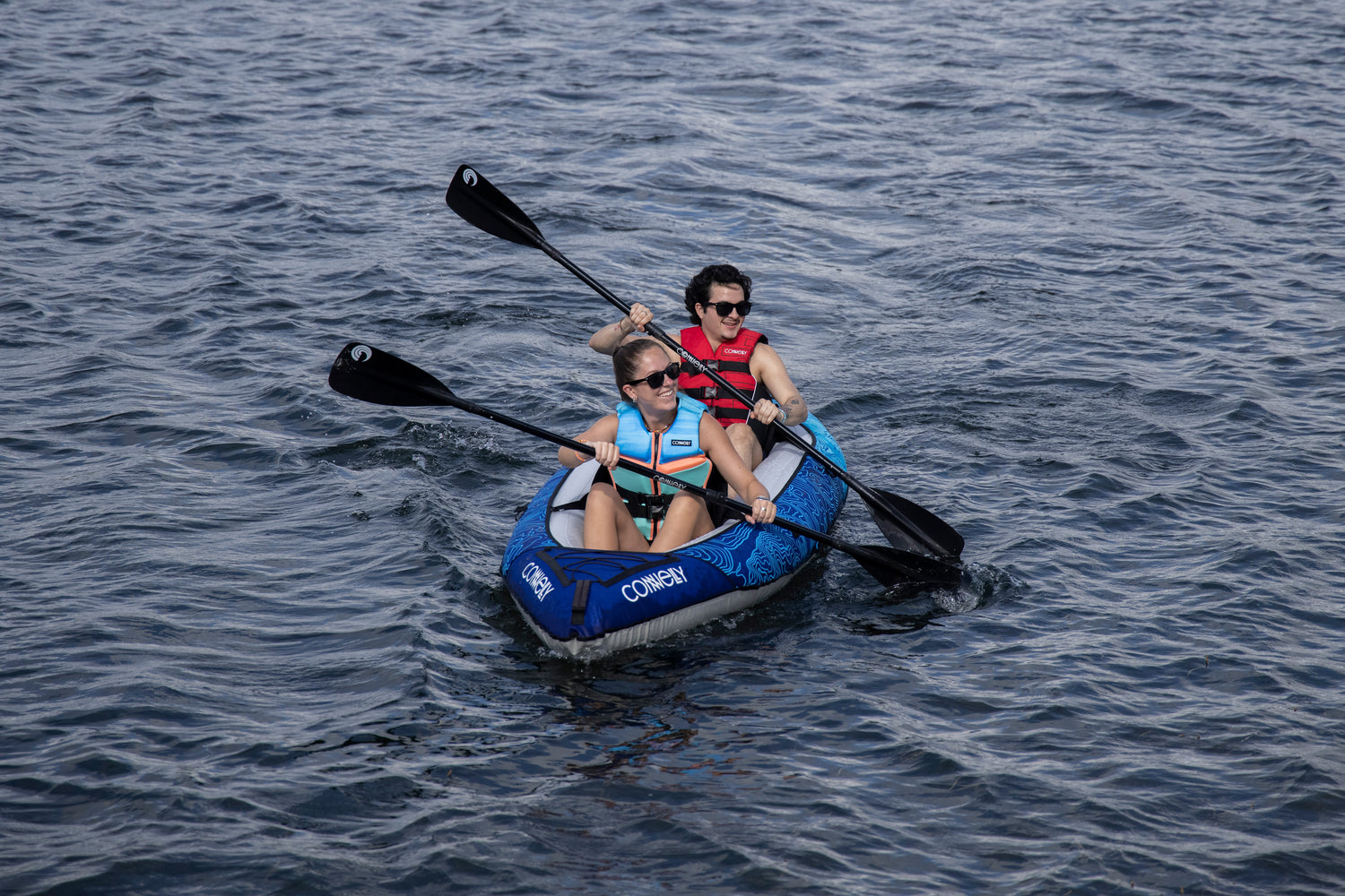 A woman and a man paddle the Nautic 11.5 inflatable kayak on a lake, both wearing life jackets and sunglasses.