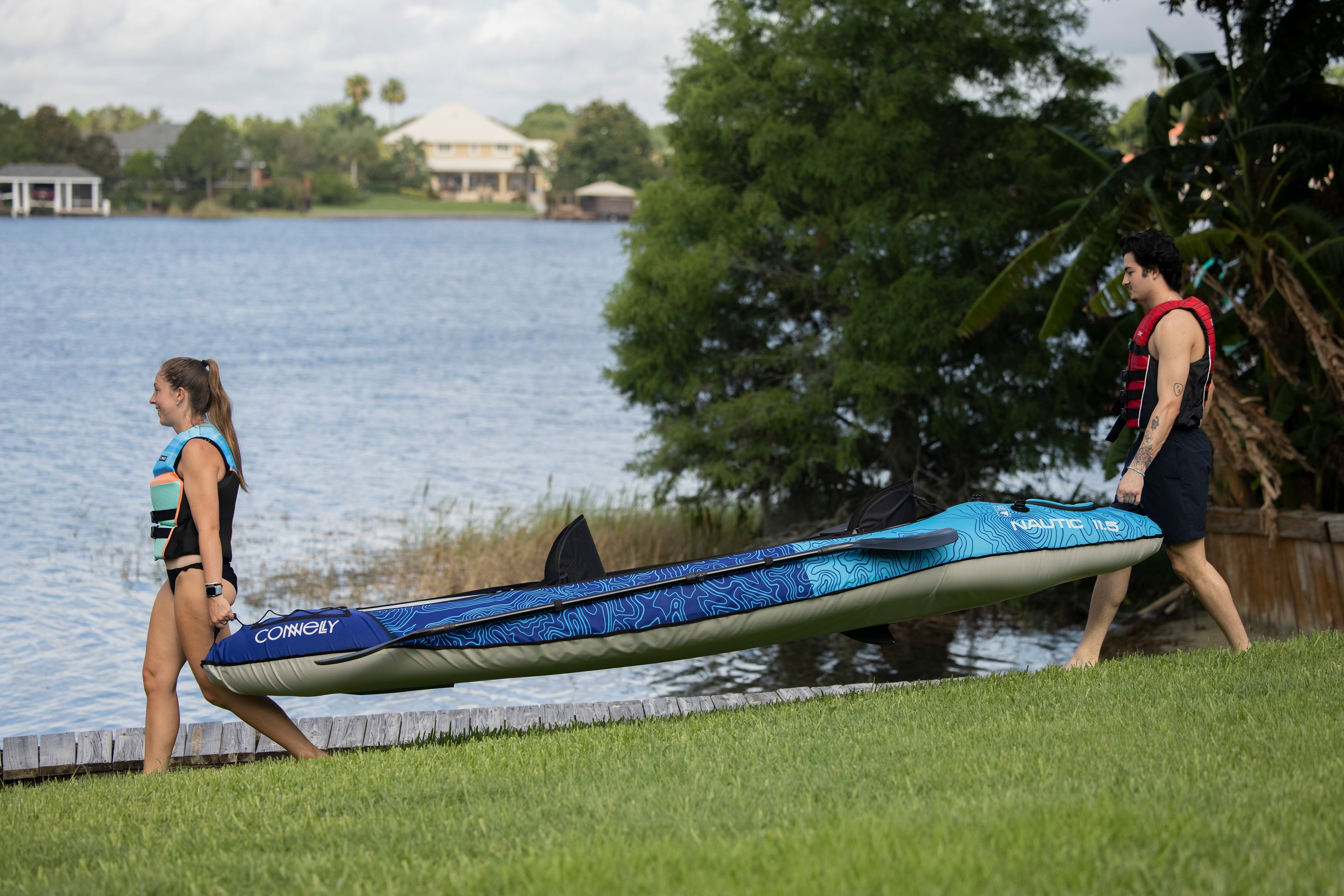 Man and woman carrying a Connelly kayak to the lake, in life vests, with scenic trees in the background.