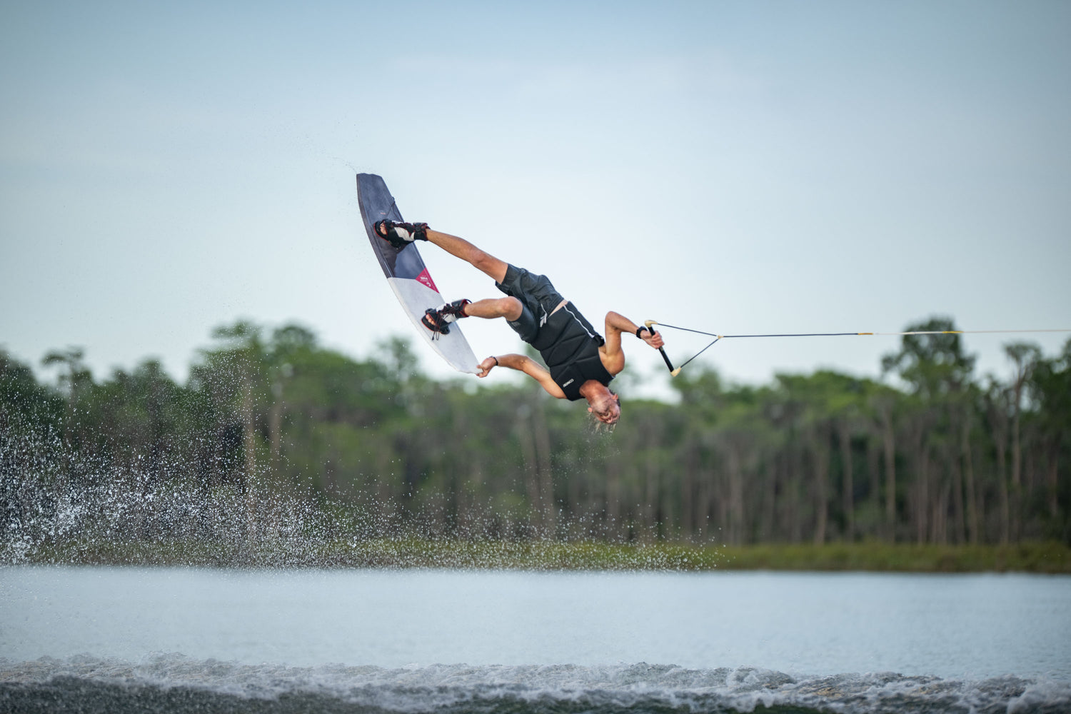 Action shot of a wakeboarder flipping mid-air while riding the PURE wakeboard on a lake.