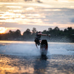 A wakeboarder carves across the water at sunset, creating a spray behind the Connelly Big Easy wakeboard.