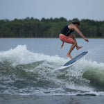 A male rider in a black vest and coral shorts performs a jump trick on the Bentley wakesurf board, mid-air above the wake.
