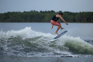 A male rider in a black vest and coral shorts performs a jump trick on the Bentley wakesurf board, mid-air above the wake.