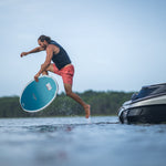 A male wakesurfer leaps off a boat’s platform onto the Connelly Bentley wakesurf board, preparing to ride the wave.