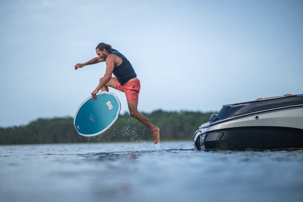 A male wakesurfer leaps off a boat’s platform onto the Connelly Bentley wakesurf board, preparing to ride the wave.
