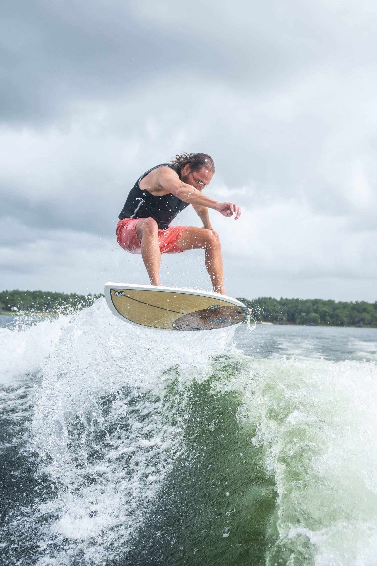 A male rider in a black vest and coral shorts carves through the wake on the Benz board.