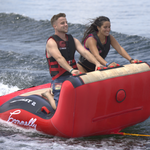 Two riders kneel and grip the front handles of the Big Easy 2 inflatable tube while riding the wake.