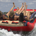 Back view of the Big Easy 3 tube in action as three riders lean back while being pulled across the water, creating a wake.