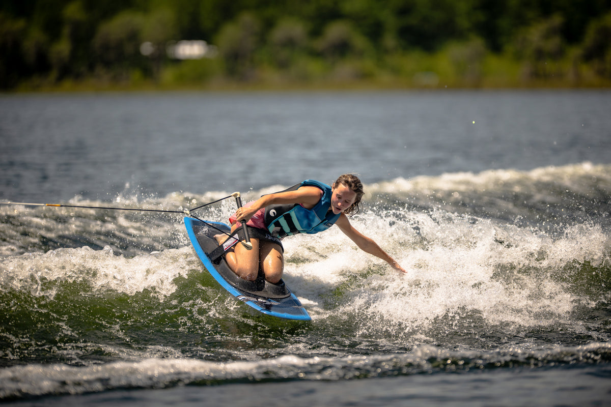 Rider wearing a blue vest carves through the water on the Boost kneeboard, gripping the handle and creating a spray.