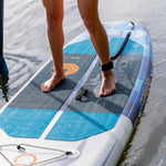 Close-up of feet standing on the Stash iSUP, highlighting the blue and black traction pad and ankle leash.