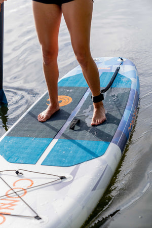 Close-up of feet standing on the Stash iSUP, highlighting the blue and black traction pad and ankle leash.