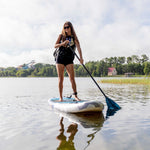 Woman paddling the Stash iSUP on a lake, wearing a black backpack, with reeds in the foreground.