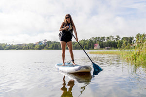 Woman paddling the Stash iSUP on a lake, wearing a black backpack, with reeds in the foreground.