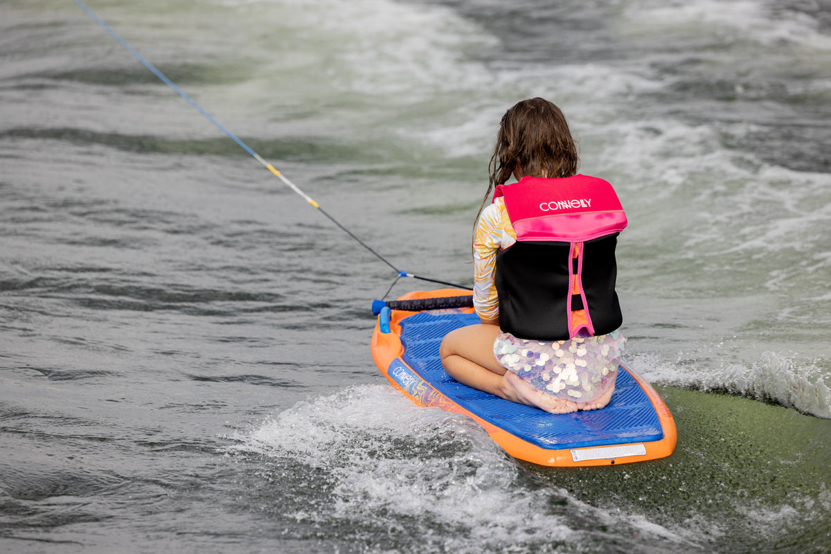 Child in pink life jacket kneeling on Wild Thing board being pulled on water.