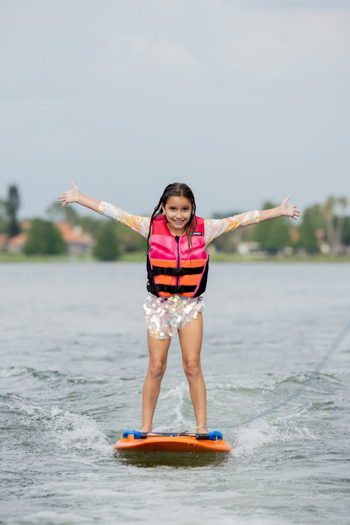 Girl on an orange wakeboard, arms out, wearing a pink Wild Thing life jacket on a lake.