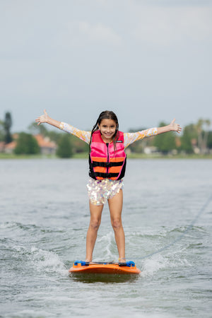Girl on an orange wakeboard, arms out, wearing a pink Wild Thing life jacket on a lake.