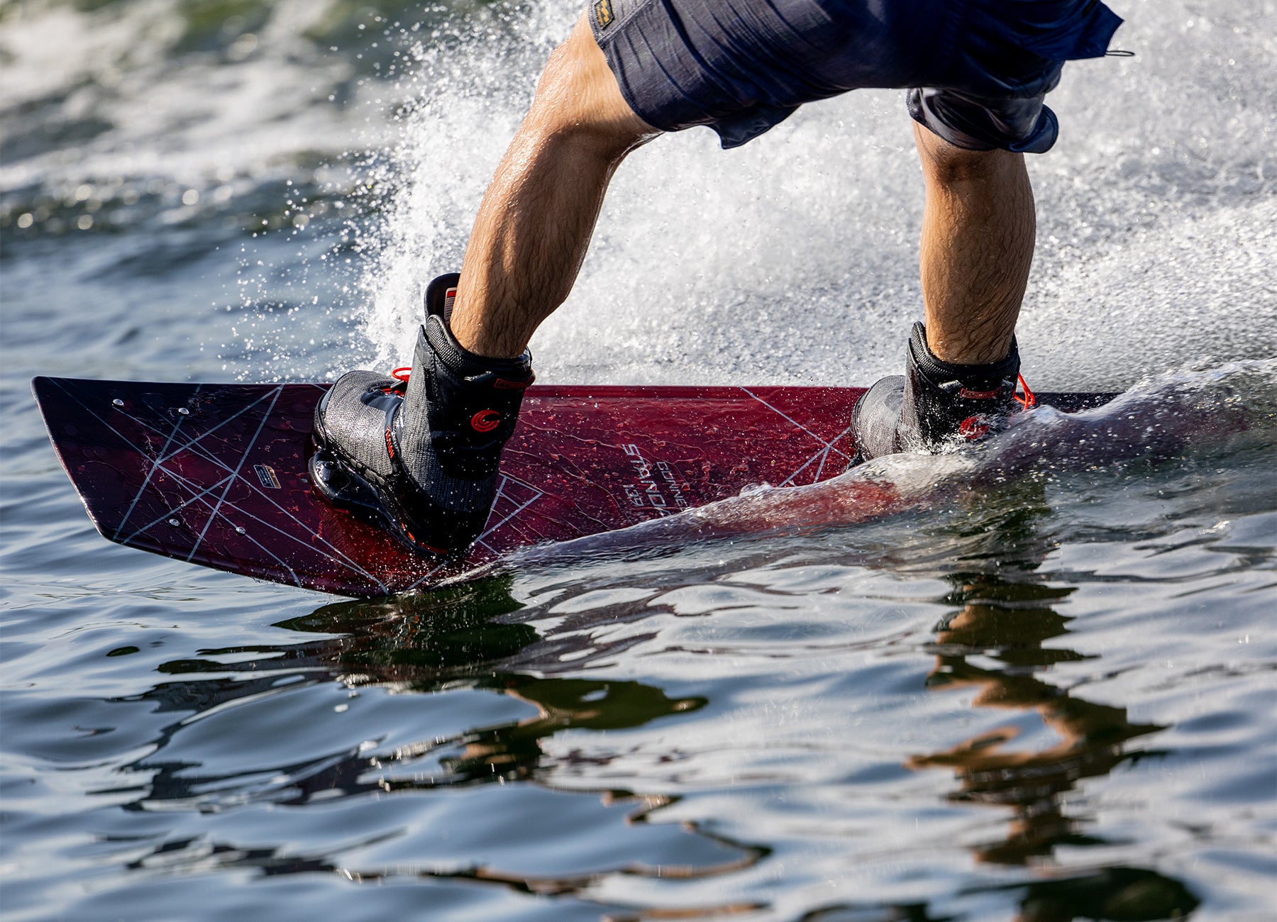 Close-up of a wakeboarder carving through the water on a red Connelly wakeboard with black bindings.