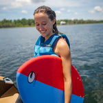 Young rider standing near the water holding the DASH Kid's Board, smiling while wearing a blue life vest.