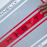 Close-up of the HABIT Skim logo on the board with red and gray stripes and a hand resting on the surface.