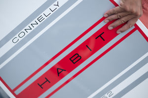 Close-up of the HABIT Skim logo on the board with red and gray stripes and a hand resting on the surface.
