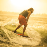 A rider carving on the HABIT Skim wakesurf board at sunset, creating a spray of water while maintaining balance.