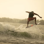 A rider mid-jump on the HABIT Skim wakesurf board, silhouetted against the water with a golden-hued sky in the background.