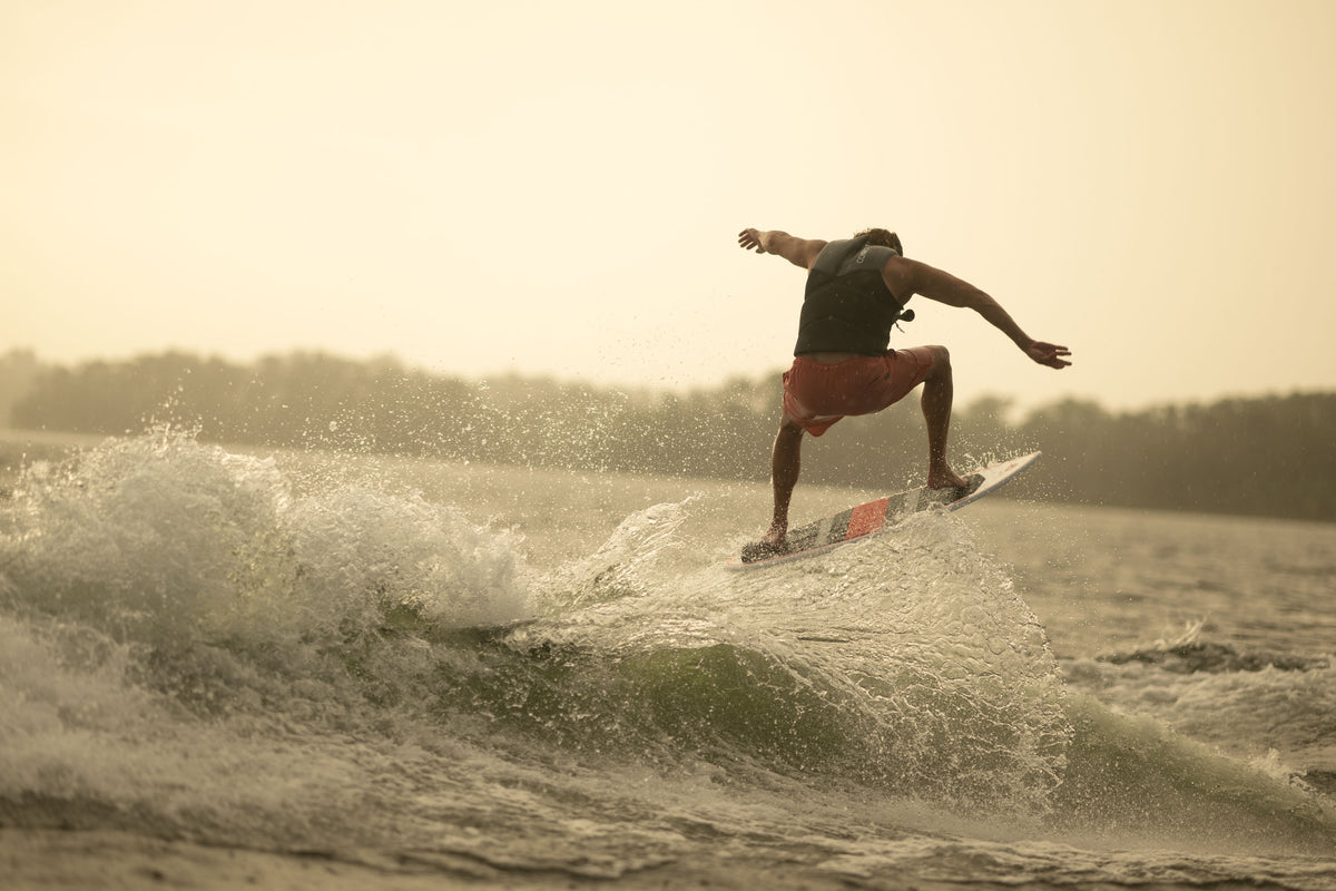 A rider mid-jump on the HABIT Skim wakesurf board, silhouetted against the water with a golden-hued sky in the background.