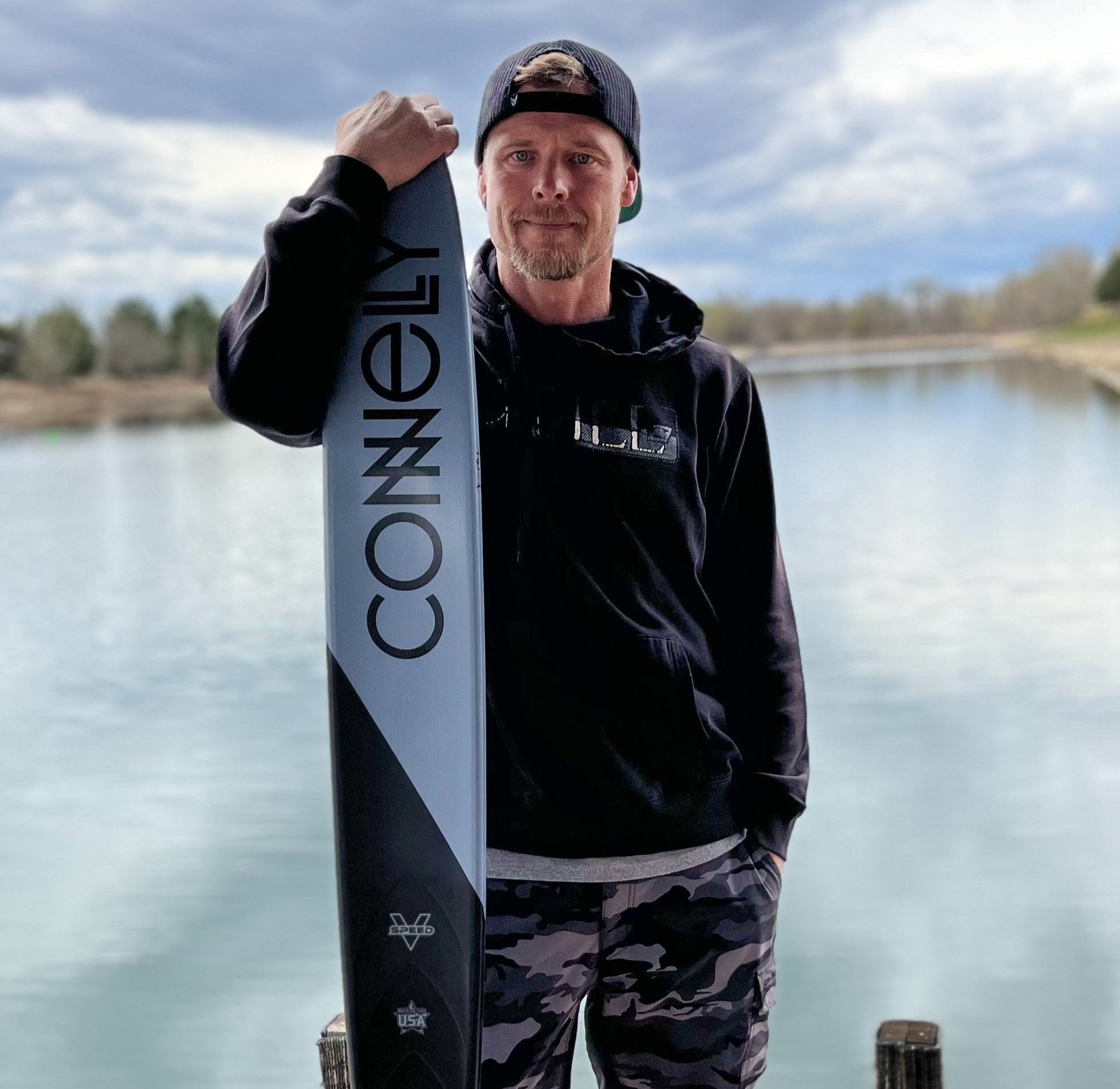 Wake skier holding a black and grey waterski brandishing the Connelly Logo wearing all black.