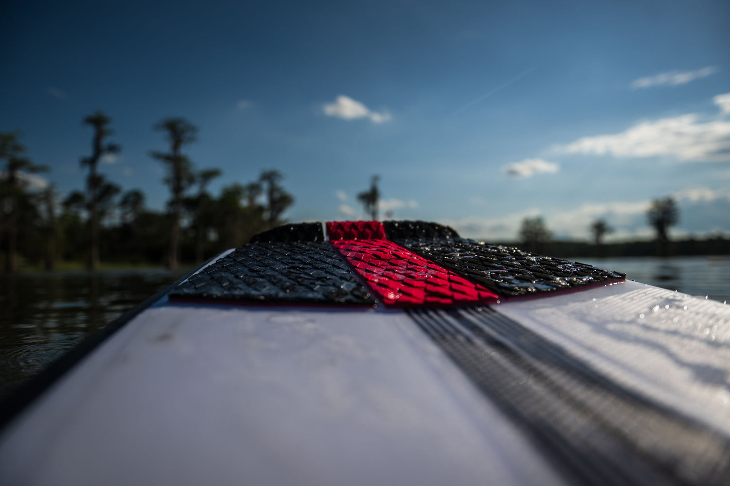 Jet wakesurf board floating on water, wet traction pad glistening under the sunlight with trees in the background.