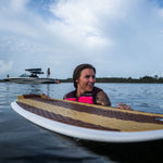 A woman in a pink life vest holds the Lil Easy wakesurf board in the water, with a boat and riders in the background.