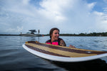 A woman in a pink life vest holds the Lil Easy wakesurf board in the water, with a boat and riders in the background.