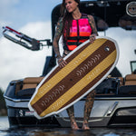 A woman in a pink life vest stands on a boat holding the Lil Easy wakesurf board, showing its textured deck and woodgrain design.