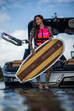 A woman in a pink life vest stands on a boat holding the Lil Easy wakesurf board, showing its textured deck and woodgrain design.