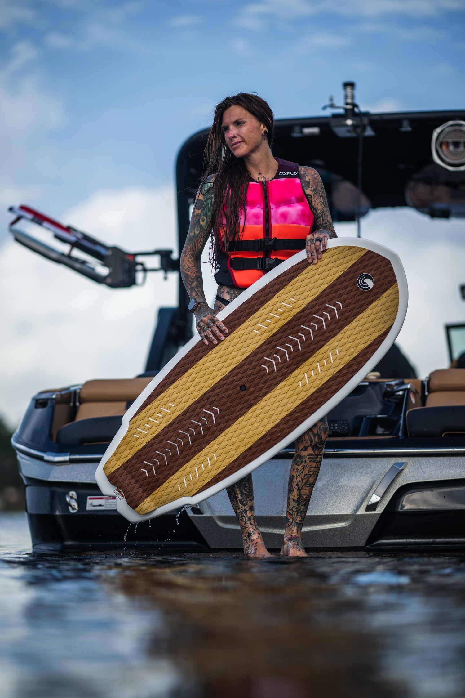 A woman in a pink life vest stands on a boat holding the Lil Easy wakesurf board, showing its textured deck and woodgrain design.