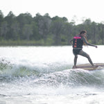 A woman in a pink life vest rides the Lil Easy wakesurf board on a wave, carving through the water.