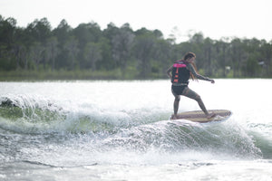 A woman in a pink life vest rides the Lil Easy wakesurf board on a wave, carving through the water.