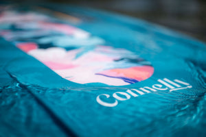 Close-up of the Lotus wakeboard’s surface, showing a blue and pink marbled graphic with the Connelly logo in water.