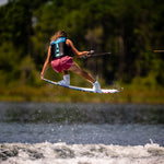 Wakeboarder mid-air on the Lotus wakeboard, wearing a black vest and pink shorts while gripping a tow rope over water.
