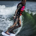 A female rider with tattoos and a pink life vest surfs a wave on the Ono wakesurf board.