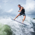 A male rider carves through a wave on the Ono wakesurf board, wearing a black vest and gray shorts.
