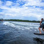 Person on a wakefoil behind a boat on a lake with trees in the background.